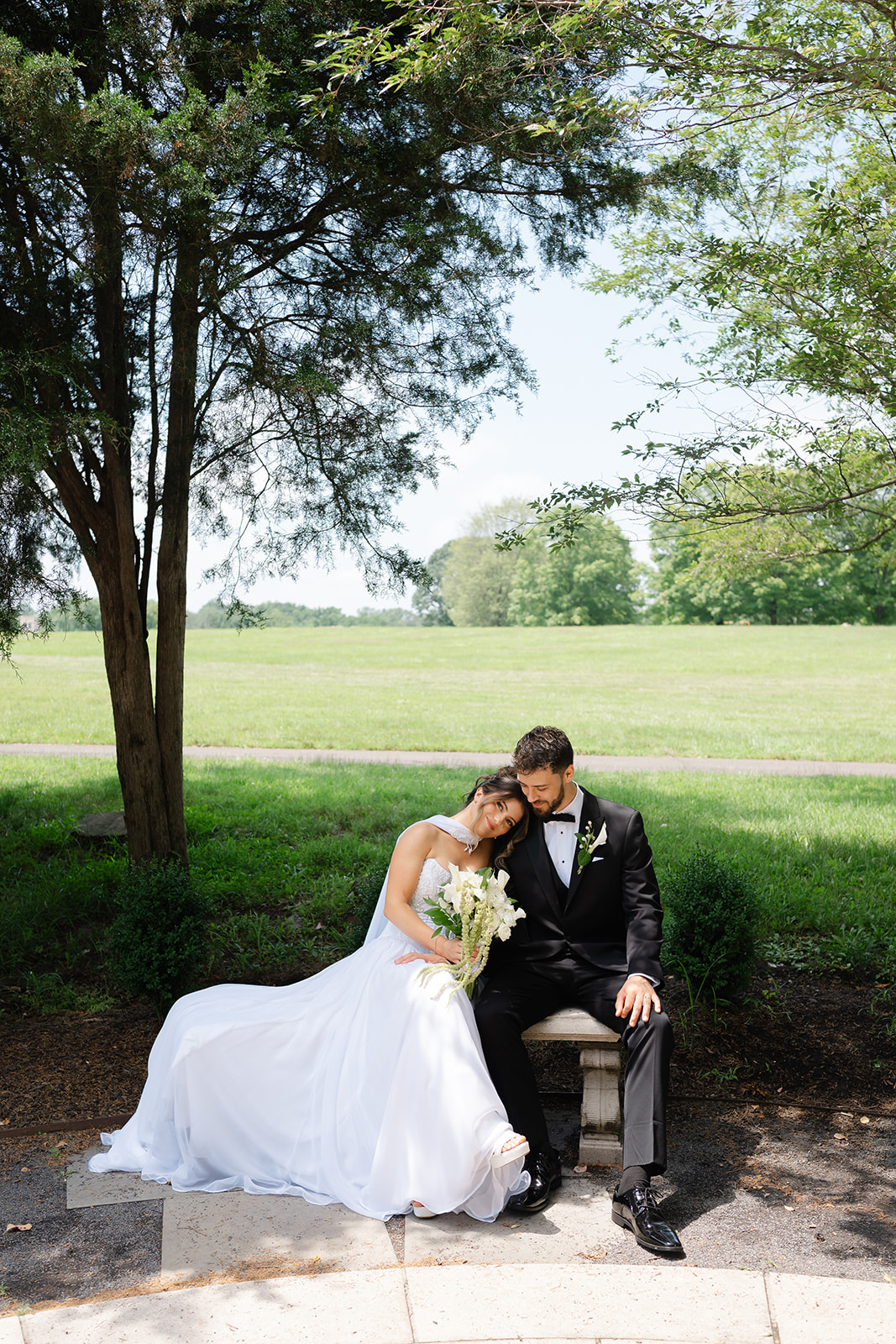 Bride and groom sitting on a stone bench under a tree, bride resting her head on groom's shoulder, holding a bouquet.