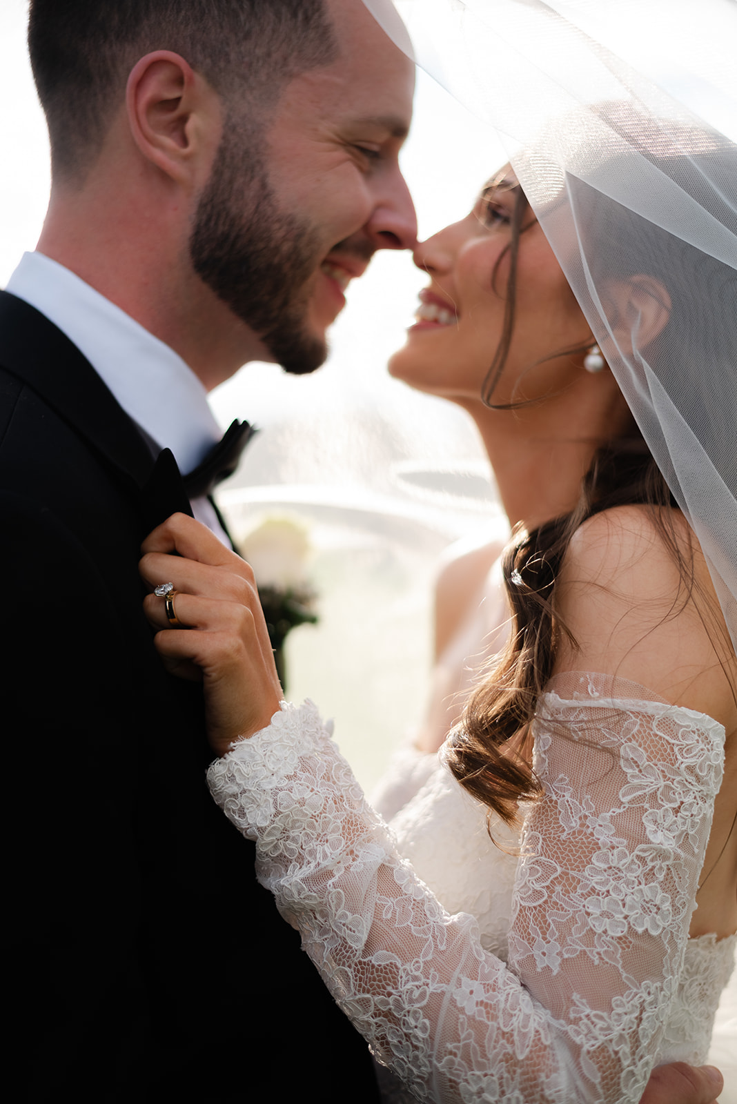 Bride and groom smiling under veil during golden hour, close-up of lace sleeve and wedding ring.